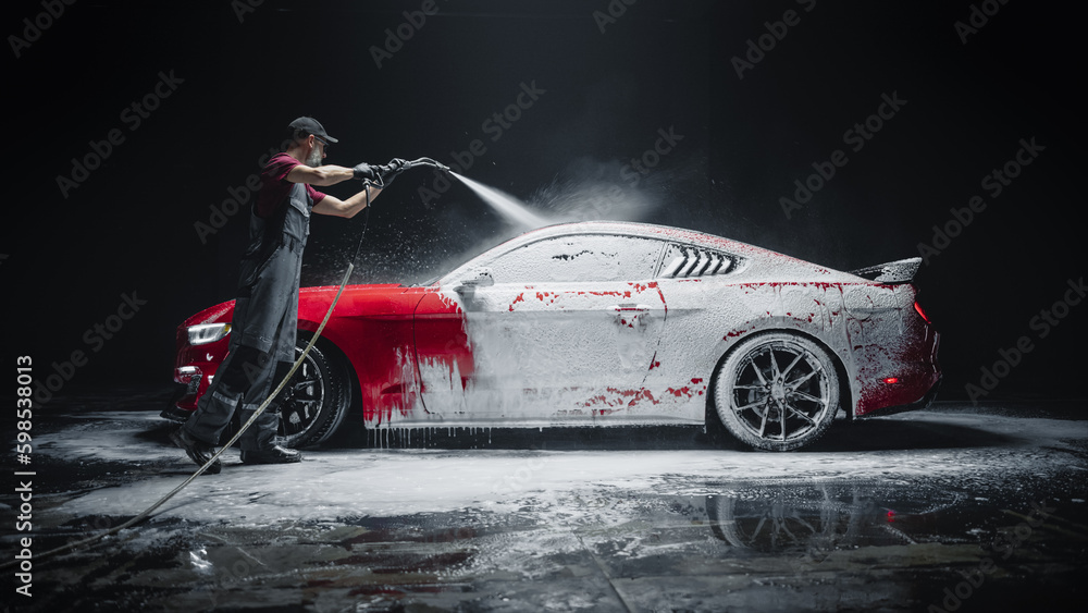 Car Wash Expert Using Water Pressure Washer to Clean a Red Modern Sportscar. Adult Man Washing Away Dirt, Preparing an American Muscle Car for Detailing. Creative Cinematic Photo with Luxury Vehicle